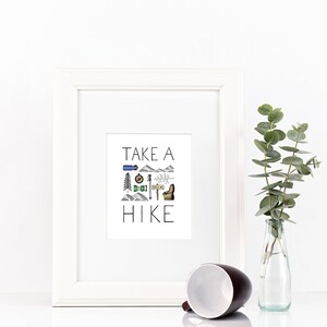 May include: A white framed print with the text "TAKE A HIKE" and hiking-themed illustrations. A brown and white mug lies on its side in front of the frame. A clear glass bottle with green leaves is to the right.