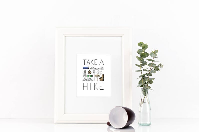 May include: A white framed print with the text "TAKE A HIKE" and hiking-themed illustrations. A brown and white mug lies on its side in front of the frame. A clear glass bottle with green leaves is to the right.