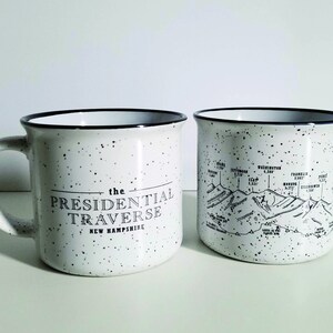 Puede incluir: Dos tazas de cer&aacute;mica moteada blanca con bordes y asas negros. Una taza presenta el texto "THE PRESIDENTIAL TRAVERSE NEW HAMPSHIRE" en negro. La otra taza muestra un mapa de cordillera en blanco y negro con nombres y elevaciones.