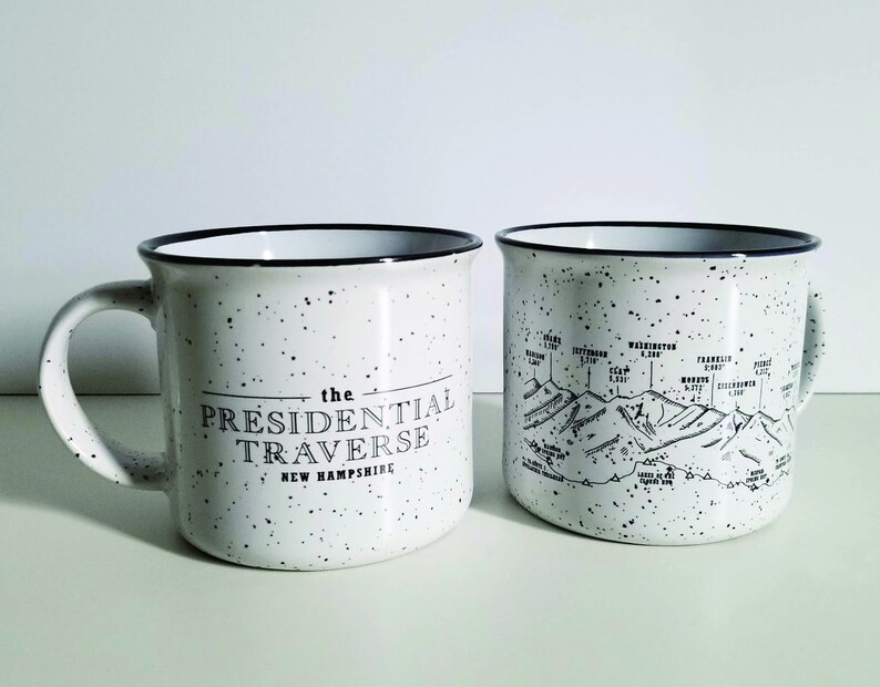 Puede incluir: Dos tazas de cer&aacute;mica moteada blanca con bordes y asas negros. Una taza presenta el texto "THE PRESIDENTIAL TRAVERSE NEW HAMPSHIRE" en negro. La otra taza muestra un mapa de cordillera en blanco y negro con nombres y elevaciones.