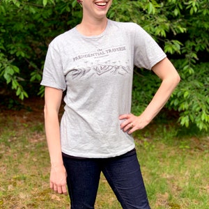 Op de afbeelding: Een vrouw draagt een grijs T-shirt met de tekst "the PRESIDENTIAL TRAVERSE NEW HAMPSHIRE" en een grafische afbeelding van een bergketen. Ze draagt ook een roze en paarse hoofdband en een blauwe jeans.