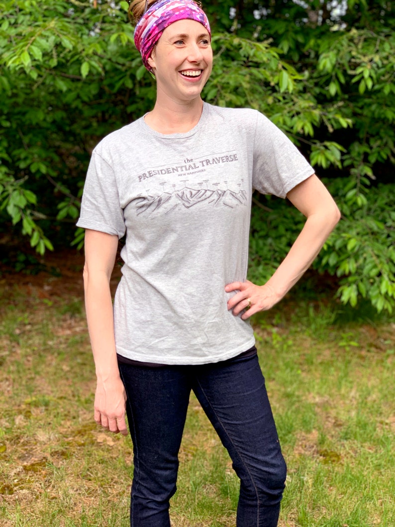Op de afbeelding: Een vrouw draagt een grijs T-shirt met de tekst "the PRESIDENTIAL TRAVERSE NEW HAMPSHIRE" en een grafische afbeelding van een bergketen. Ze draagt ook een roze en paarse hoofdband en een blauwe jeans.