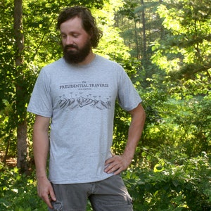 Op de afbeelding: Een man draagt een grijs T-shirt met de tekst "The Presidential Traverse New Hampshire" erop gedrukt. Hij staat in een bos.