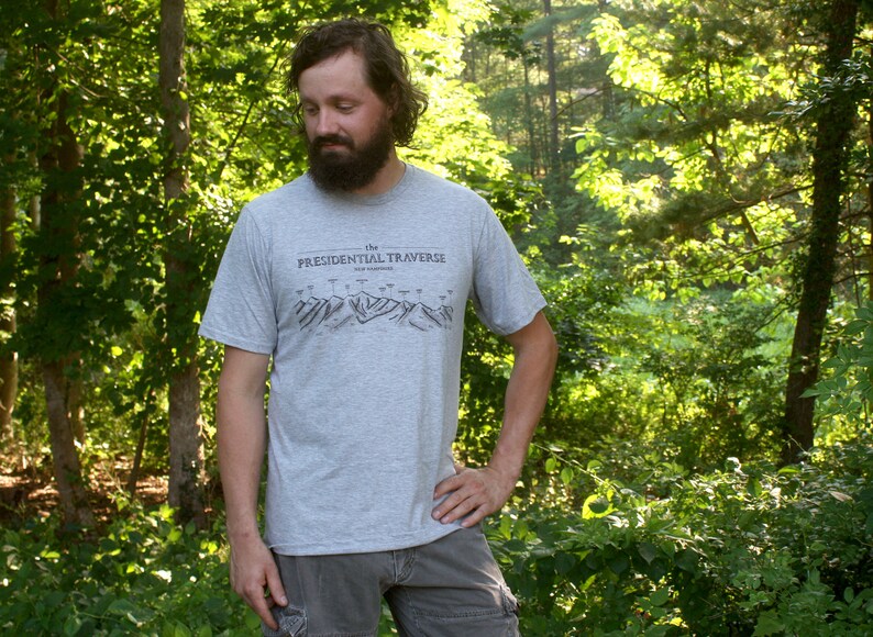 Op de afbeelding: Een man draagt een grijs T-shirt met de tekst "The Presidential Traverse New Hampshire" erop gedrukt. Hij staat in een bos.