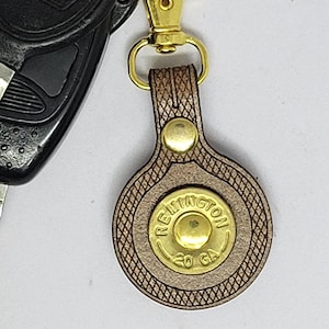 May include: A collection of car keys and a keychain. The keychain has a brown leather backing, a gold-coloured metal clasp, and a bullet casing design. The bullet casing is embossed with the word "Remington" and "20 GA".