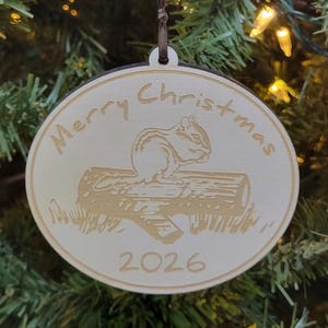 May include: A round, light-colored wooden Christmas ornament hanging from a dark brown ribbon. The ornament features an engraved image of a chipmunk sitting on a log, with the text Merry Christmas above and the year 2026 printed below the image.