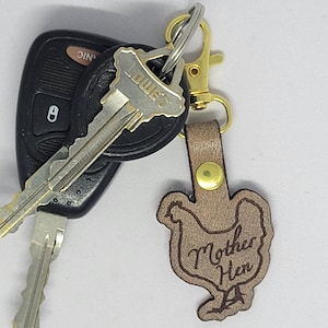 May include: A tan leather keychain with a hen silhouette and the words "Mother Hen" engraved on it. The keychain is attached to a gold-colored clasp and a set of car keys.