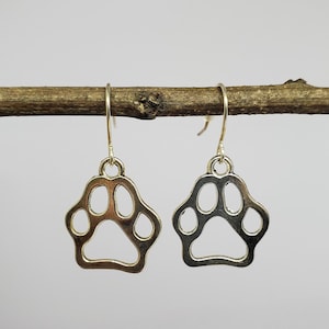 May include: A pair of gold-tone dangle earrings featuring a paw print design. The earrings are hanging from a branch.