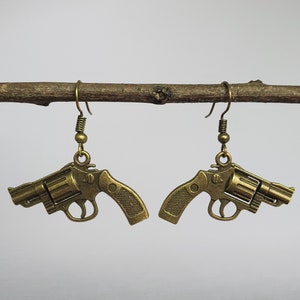 May include: A pair of gold-toned earrings shaped like revolvers. The earrings are hanging from a branch.