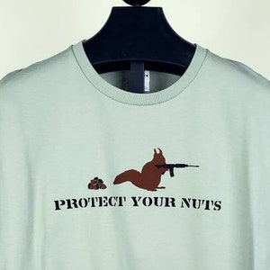 May include: A light green t-shirt with a brown squirrel holding a rifle and a pile of nuts. The text "PROTECT YOUR NUTS" is printed below the squirrel.