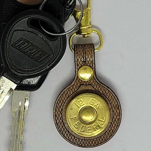 May include: A keychain with a brown leather fob and a brass-colored metal accent. The fob is shaped like a bullet and has the text "12 GA FEDERAL". Attached to the fob are keys and a car key.
