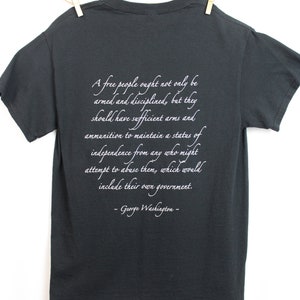 Puede incluir: Una camiseta gris oscura con una cita blanca de George Washington sobre la importancia de un pueblo libre armado y disciplinado. La cita dice: "Un pueblo libre no solo debe estar armado y disciplinado, sino que debe tener suficientes armas y municiones para mantener un estado de independencia de cualquiera que pueda intentar abusar de ellos, lo que incluir&iacute;a su propio gobierno. -George Washington- "