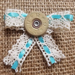 May include: A white lace bow with turquoise ribbon and a gold shotgun shell in the center. The shell has the numbers "12" and "12" printed on it, and the words "12 Gauge" are printed on the side.
