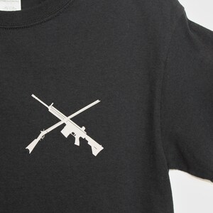 Puede incluir: Camiseta negra con una silueta blanca de dos rifles cruzados.
