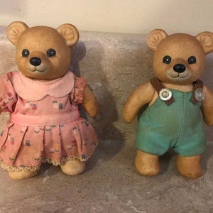 May include: Two ceramic teddy bears, one wearing a pink dress with floral print and the other wearing blue overalls. Both bears have brown fur and black eyes.