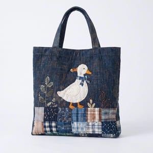 Puede incluir: Un bolso tote de mezclilla azul oscuro con un pato blanco bordado con pico y patas naranjas, que lleva una bufanda de cuadros azules. La parte inferior del bolso está decorada con un patchwork de varios cuadrados de tela a cuadros y estampados.