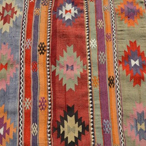 May include: Close-up of a colorful woven rug with vertical stripes in shades of red, orange, purple, and gray. The rug features geometric patterns in various colors, including white, pink, and black. The rug is likely made of wool or a similar material.