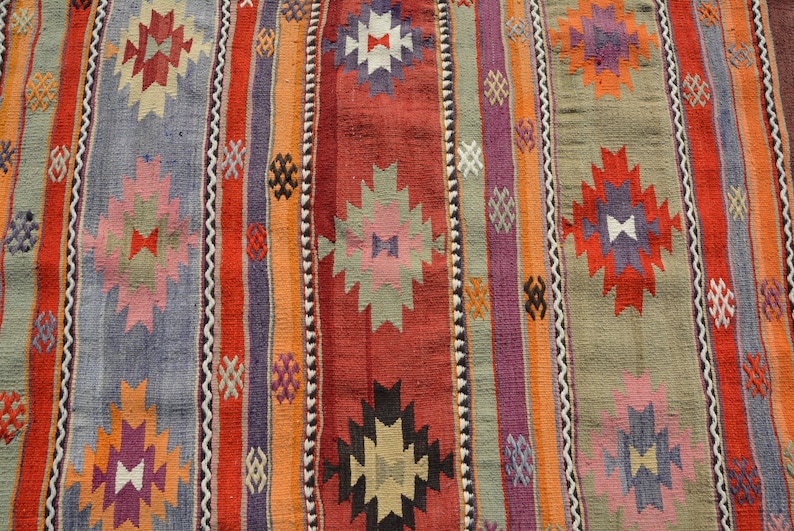 May include: Close-up of a colorful woven rug with vertical stripes in shades of red, orange, purple, and gray. The rug features geometric patterns in various colors, including white, pink, and black. The rug is likely made of wool or a similar material.