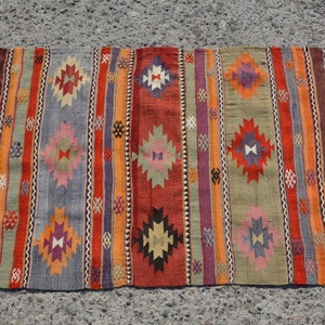 May include: A rectangular, hand-woven rug with a striped pattern in shades of red, orange, gray, and green. The rug features geometric diamond shapes and fringe on the short ends. The rug is approximately 36 inches long and 24 inches wide.