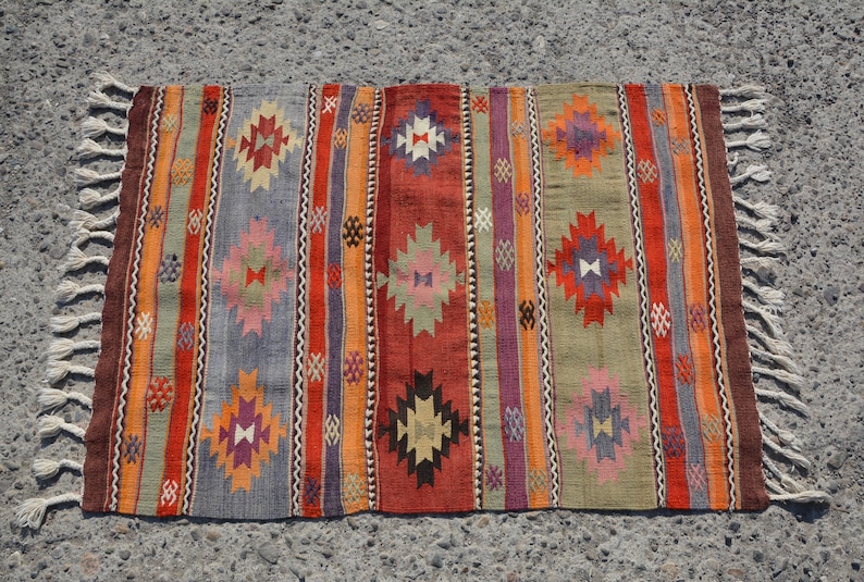 May include: A rectangular, hand-woven rug with a striped pattern in shades of red, orange, gray, and green. The rug features geometric diamond shapes and fringe on the short ends. The rug is approximately 36 inches long and 24 inches wide.