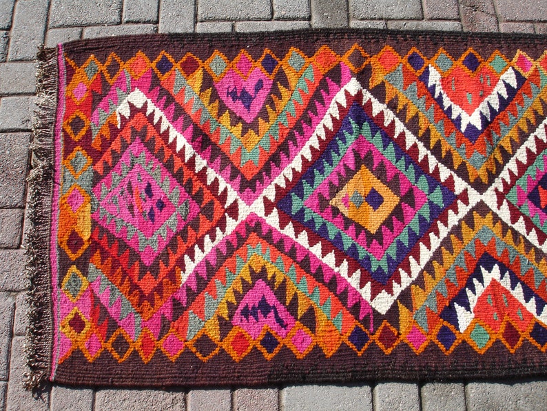 May include: A colorful rug with a geometric pattern in shades of red, orange, pink, purple, green, and white. The rug has a brown border and fringe.
