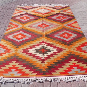 May include: A colorful rug with a geometric pattern in shades of red, orange, yellow, brown, and gray. The rug has a fringe border.