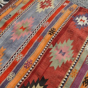May include: A colorful, handwoven rug with a striped pattern. The rug features a variety of colors, including red, orange, purple, and gray. Geometric designs are incorporated throughout the rug's design. The rug has a fringed edge.
