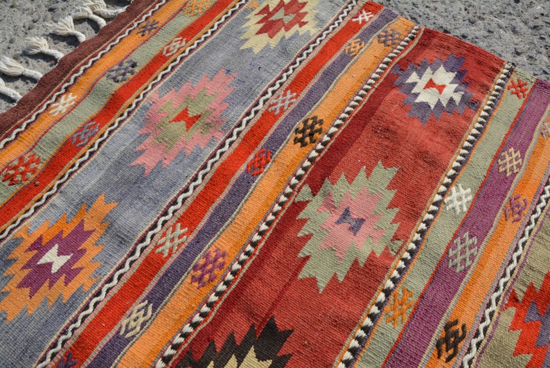 May include: A colorful, handwoven rug with a striped pattern. The rug features a variety of colors, including red, orange, purple, and gray. Geometric designs are incorporated throughout the rug's design. The rug has a fringed edge.