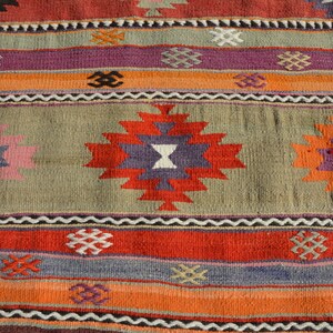 May include: Close-up of a colorful woven rug with geometric patterns. The rug features a central design with red, purple, and white shapes, surrounded by stripes of orange, red, and gray. The rug has a textured appearance.