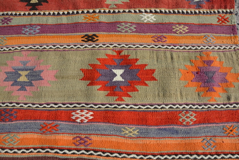 May include: Close-up of a colorful woven rug with geometric patterns. The rug features a central design with red, purple, and white shapes, surrounded by stripes of orange, red, and gray. The rug has a textured appearance.
