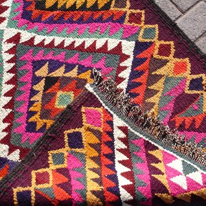 May include: A colorful handwoven rug with geometric patterns in shades of pink, red, yellow, green, and brown. The rug has a fringe border and is made of wool.