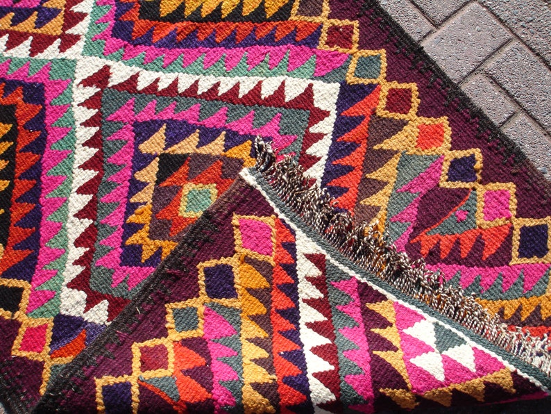 May include: A colorful handwoven rug with geometric patterns in shades of pink, red, yellow, green, and brown. The rug has a fringe border and is made of wool.