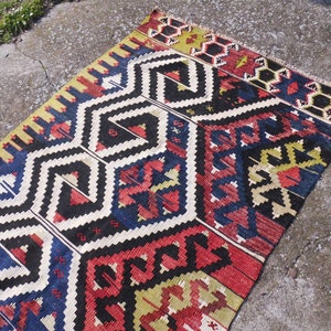 May include: A colorful, geometric patterned rug with a black background and red, blue, yellow, and white accents. The rug has a traditional design and is made of wool.