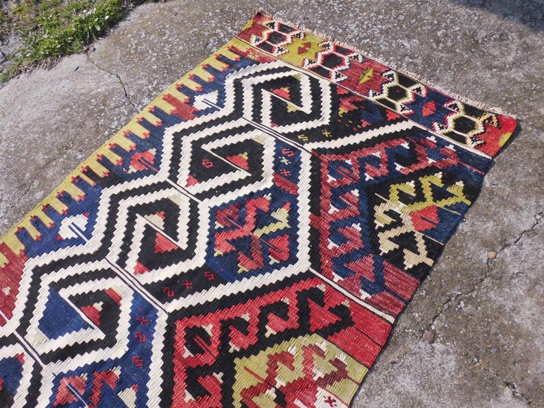 May include: A colorful, geometric patterned rug with a black background and red, blue, yellow, and white accents. The rug has a traditional design and is made of wool.
