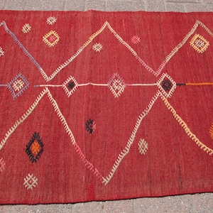 May include: A red woven rug with geometric patterns. The rug features large white zigzags and diamond shapes in various colors, including orange, yellow, and black. The rug is made of a textured material.