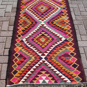 May include: A colorful, geometric patterned rug with a diamond design in shades of pink, orange, yellow, green, and purple. The rug has a dark brown border and fringe.