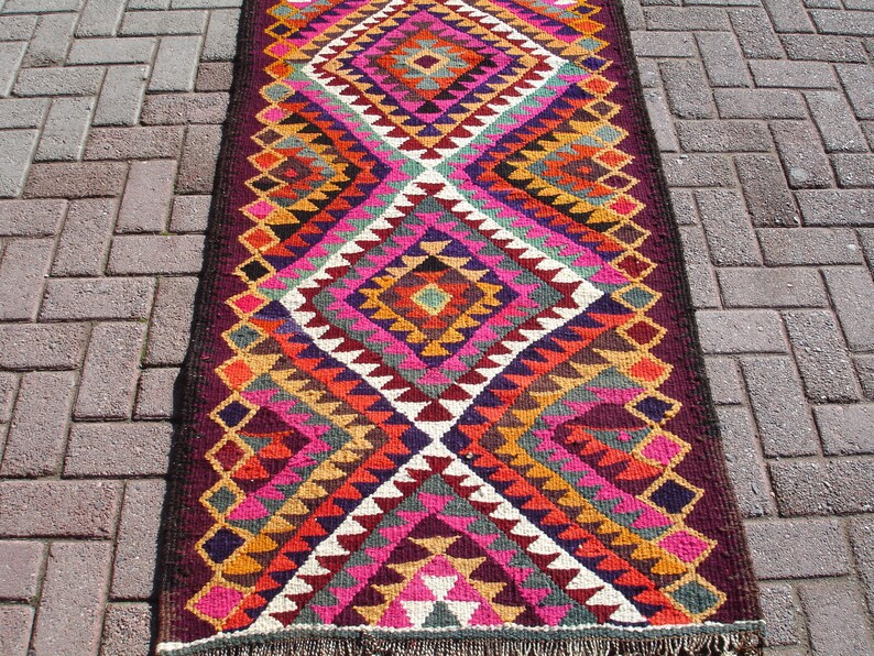 May include: A colorful, geometric patterned rug with a diamond design in shades of pink, orange, yellow, green, and purple. The rug has a dark brown border and fringe.