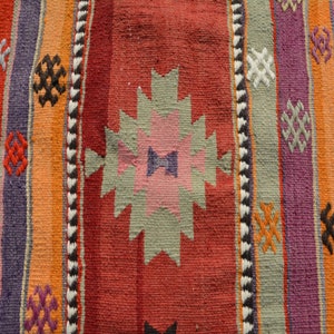 May include: Close-up of a woven rug with a colorful striped pattern. The rug features vertical stripes in shades of red, orange, purple, and blue, with geometric designs and a zig-zag border. The rug's texture is visible.