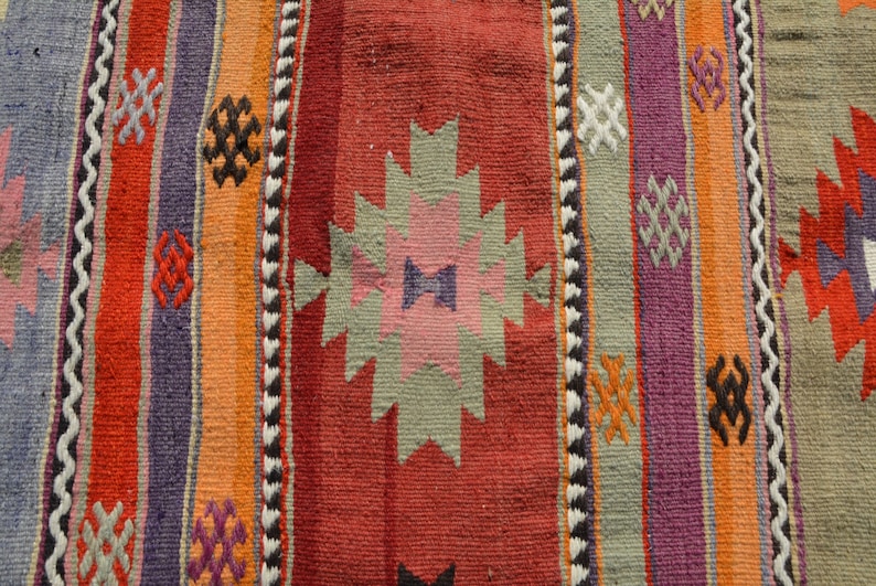 May include: Close-up of a woven rug with a colorful striped pattern. The rug features vertical stripes in shades of red, orange, purple, and blue, with geometric designs and a zig-zag border. The rug's texture is visible.