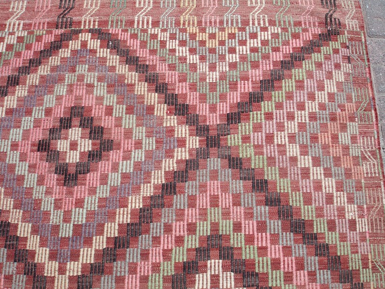 K&ouml;nnte beinhalten: Ein Vintage-Teppich mit einem geometrischen Muster in Rot-, Rosa-, Gr&uuml;n- und Schwarzt&ouml;nen. Der Teppich hat eine gewebte Textur und einen abgenutzten Look.