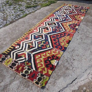 May include: A long, narrow rug with a geometric pattern in shades of red, yellow, blue, and black. The rug has a fringed edge and is made of wool.