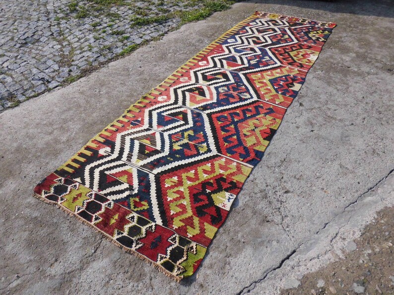 May include: A long, narrow rug with a geometric pattern in shades of red, yellow, blue, and black. The rug has a fringed edge and is made of wool.