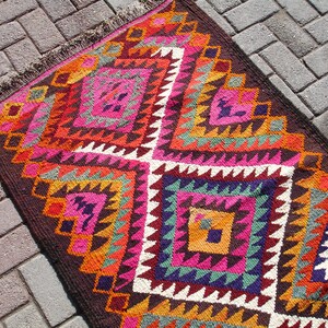 May include: A colorful rug with a geometric pattern in shades of pink, orange, brown, green, and white. The rug has a fringe border and is woven with a traditional design.