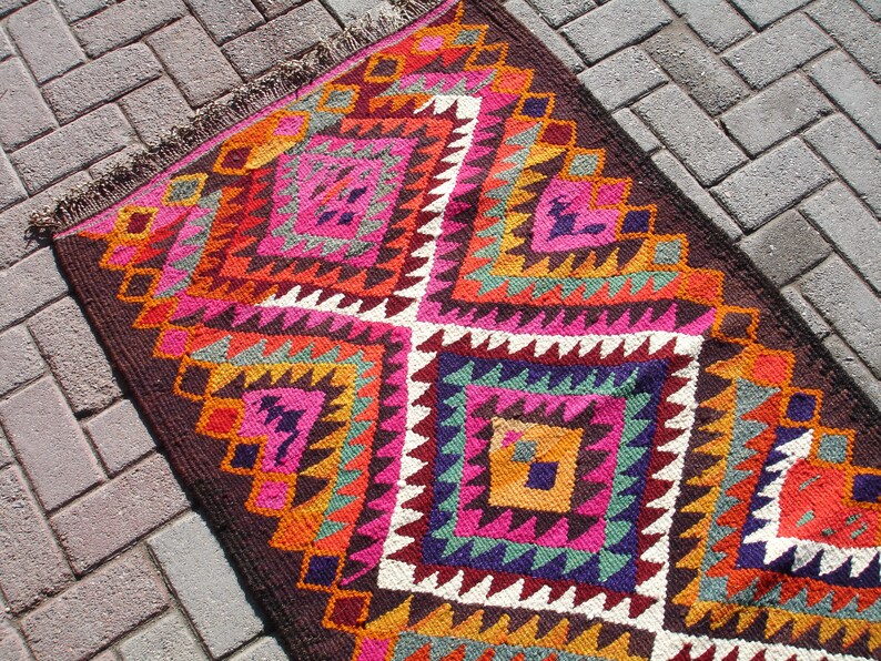 May include: A colorful rug with a geometric pattern in shades of pink, orange, brown, green, and white. The rug has a fringe border and is woven with a traditional design.