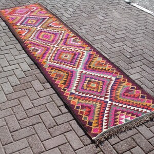 May include: A long, narrow rug with a geometric pattern in shades of pink, orange, yellow, and brown. The rug has a fringe on both ends.