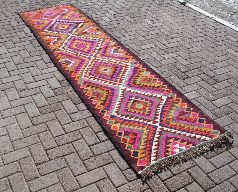 May include: A long, narrow rug with a geometric pattern in shades of pink, orange, yellow, and brown. The rug has a fringe on both ends.
