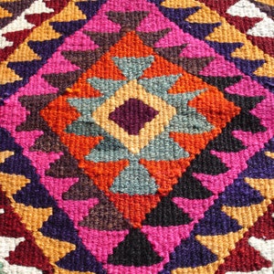 May include: A colorful rug with a geometric pattern in shades of red, orange, pink, purple, blue, and white. The rug is woven with a diamond-shaped design in the center.
