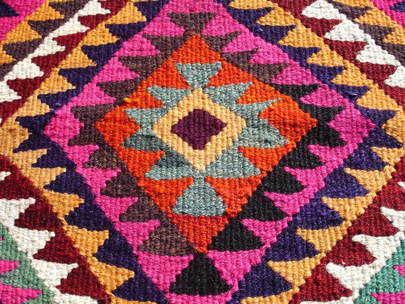 May include: A colorful rug with a geometric pattern in shades of red, orange, pink, purple, blue, and white. The rug is woven with a diamond-shaped design in the center.