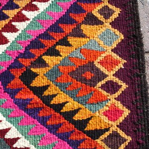 May include: A colorful rug with a geometric pattern in shades of red, orange, yellow, green, blue, purple, and white. The rug has a fringe border.