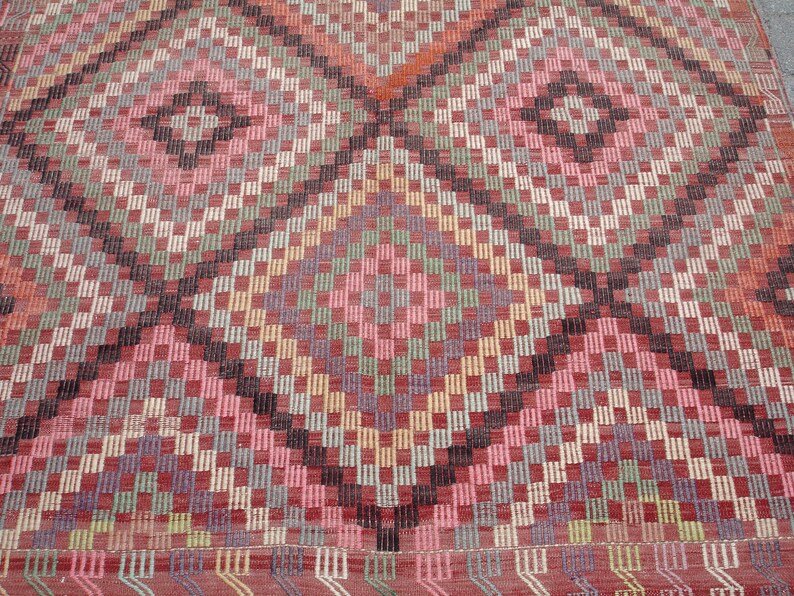 K&ouml;nnte beinhalten: Ein bunter Teppich mit einem geometrischen Muster in Rot-, Rosa-, Gr&uuml;n-, Blau- und Schwarzt&ouml;nen. Der Teppich hat eine Diamantform in der Mitte und ist mit verschiedenen Texturen gewebt.